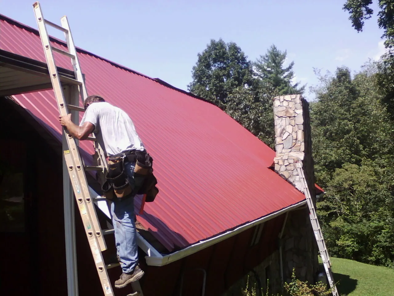 Roofer on a ladder beside a red metal roof and stone chimney — Soffit & Fascia Repair by Roofing Brothers in Barrett